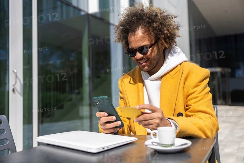 Afro man in a stylish yellow coat and sunglasses is sitting at an outdoor cafe table, smiling while using his smartphone and a credit card, portraying a relaxed moment