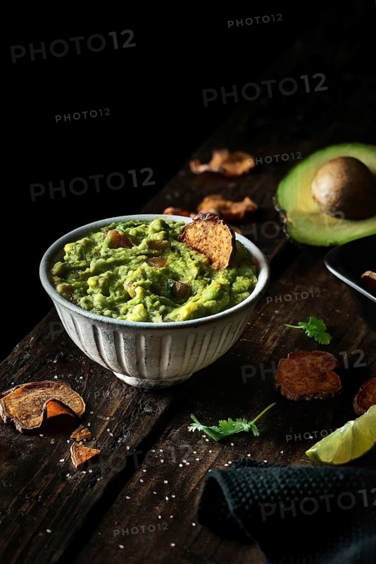 Top view of bowl of fresh guacamole placed near avocado and chips on weathered lumber tabletop
