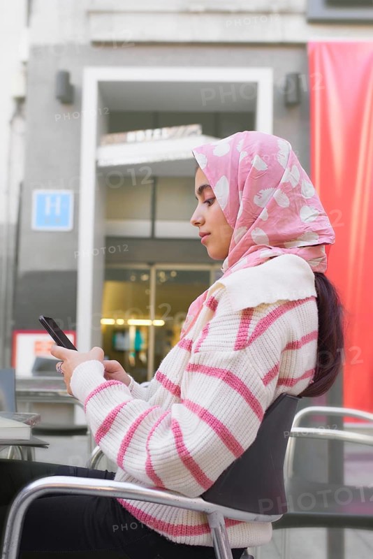 A arab student woman in a striped sweater sits at an outdoor cafe, focused on her smartphone. A modern, urban background complements the serene, everyday moment captured