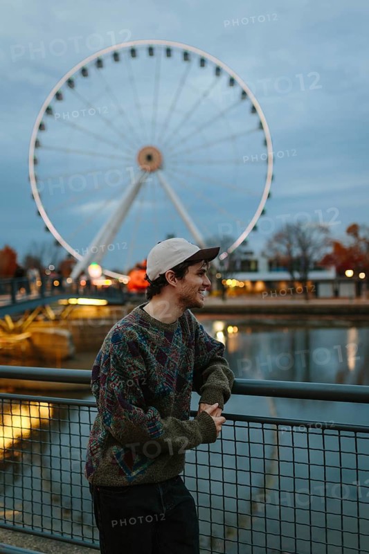 Side view of man in a colorful sweater leans on a railing, smiling as the iconic Montreal Ferris wheel illuminates the background on a crisp autumn evening