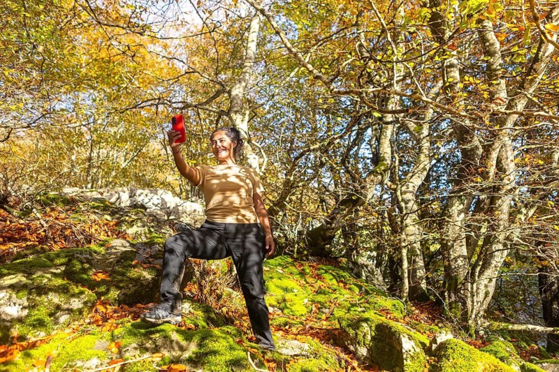 A woman enjoys a scenic autumn hike in Gorbea Natural Park, Basque Country, Spain. Surrounded by vibrant foliage, she captures with a selfie the beauty of nature with her phone