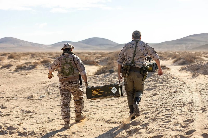 Two soldiers, dressed in camouflage, carry a heavy supply box across an arid desert landscape, showcasing teamwork and military operation in harsh environments