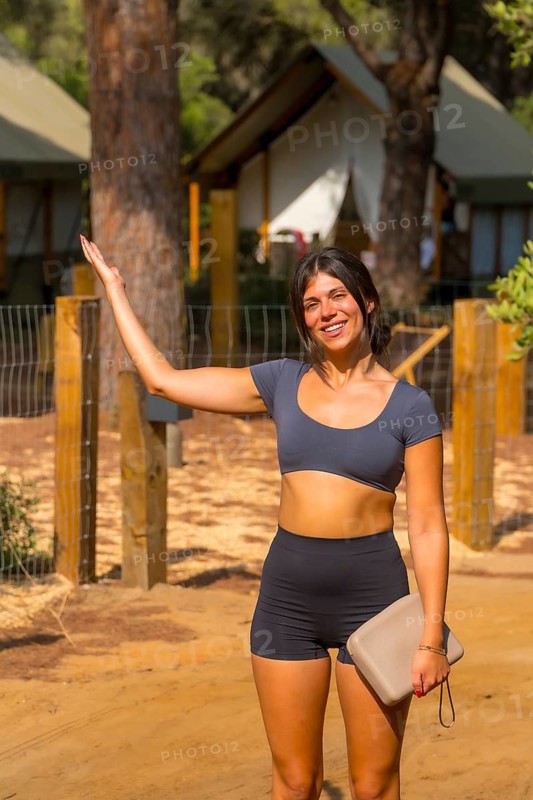 Young tourist woman with a clutch bag waving hand and smiling at the entrance of a glamping resort, enjoying her vacation surrounded by nature and comfortable accommodations
