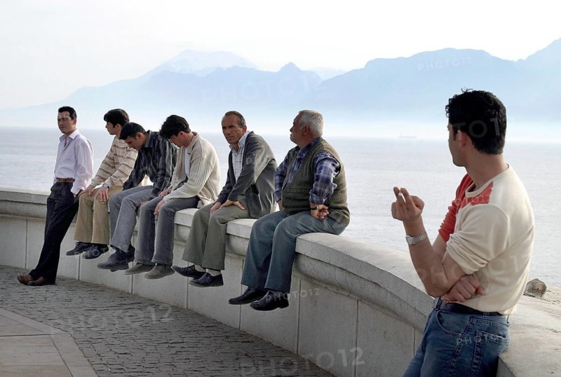 Antalya, TUR, 10.04.2004 - Turkish men sit on a wall on the waterfront in Antalya, Turkey. In the background the Taurus Mountains, Turkey