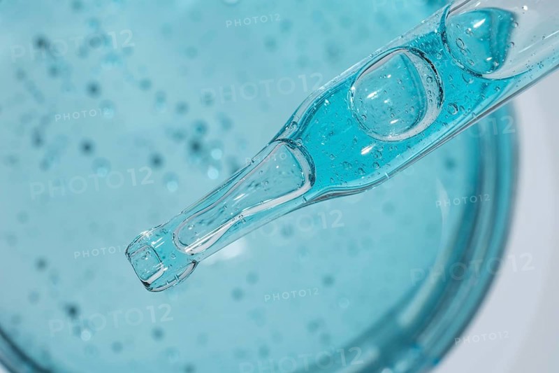 Close-up of blue cosmetic gel with visible droplets and texture, captured from above. A clear pipette hovers over the gel, highlighting bubble formations