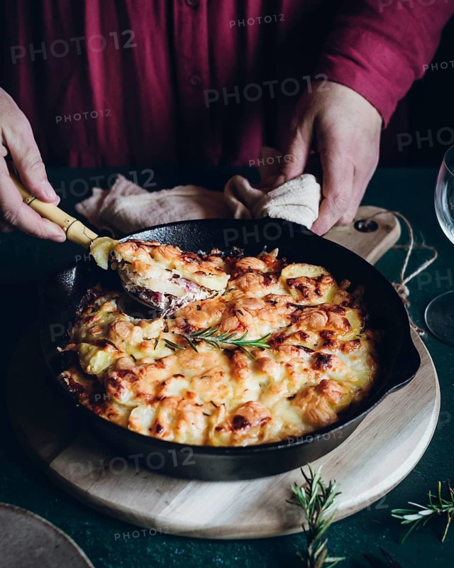 A homemade potato gratin casserole baked to golden perfection in a cast iron skillet. A person serves a portion on a cozy table, creating a rustic and inviting dining atmosphere
