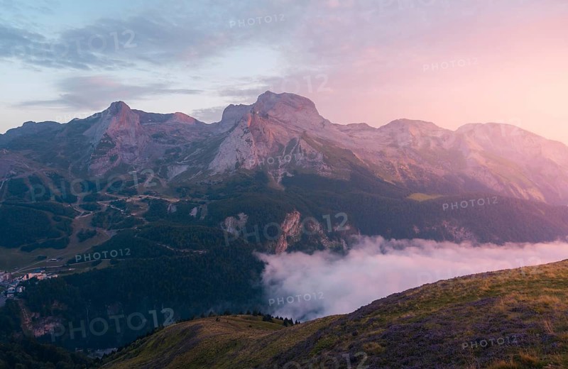 A stunning mountain landscape captured during dawn, with warm pink hues illuminating the rocky peaks in the Pyrenees. Misty clouds drift below, enhancing the serene atmosphere in summer