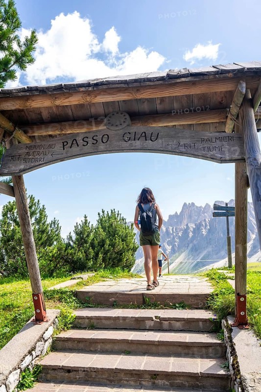 Tourists walking under a wooden sign indicating the giau pass in the italian dolomites, with a stunning mountain view in the background