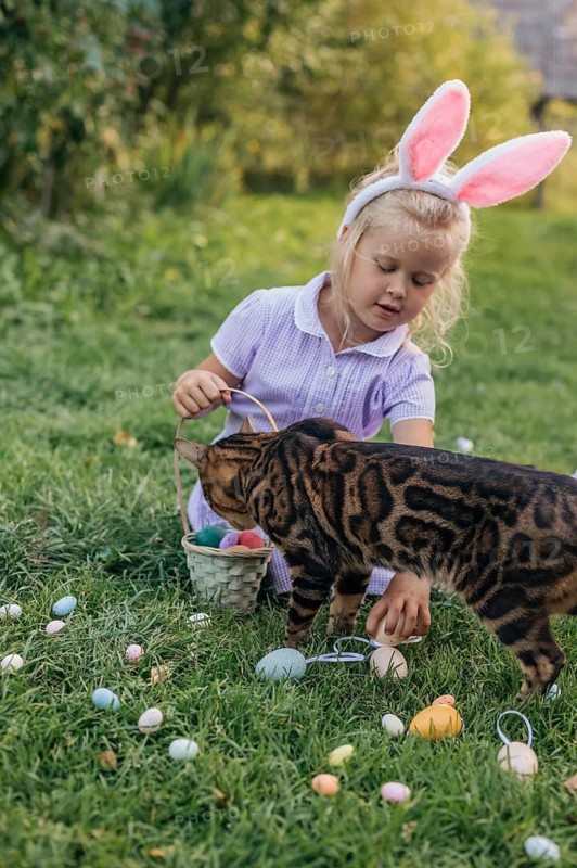 A young girl wearing bunny ears is sitting on a grassy field, placing colorful Easter eggs into a basket. A curious cat joins in the festive moment