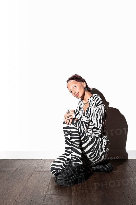 A fashion model poses in a striking black-and-white zebra print outfit, sitting under bright studio lighting. Flash photography highlights her unique style and expression
