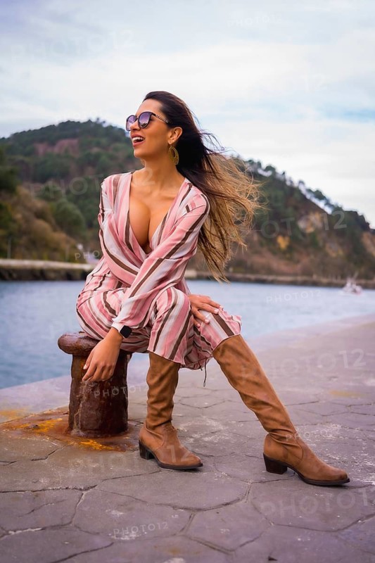 Portrait of a young Latin woman in a pink dress, brown boots and sunglasses enjoying summer vacations, sitting by the sea