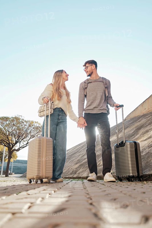 A young couple walks hand in hand with suitcases, exploring Majorca, Spain. They're on a sunny adventure, excited to find their dream vacation home near Calatrava