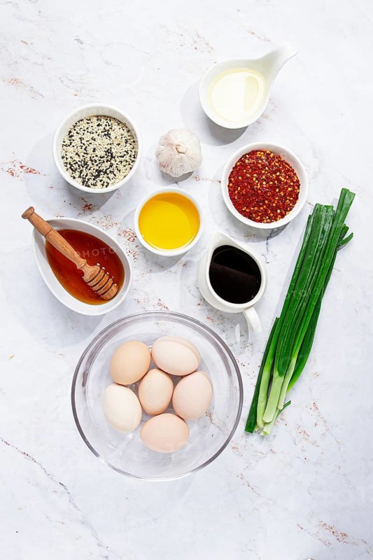 The countertop shows several bowls with ingredients for a dish. There are eggs, garlic, green onions, oils, honey, and spices ready for cooking