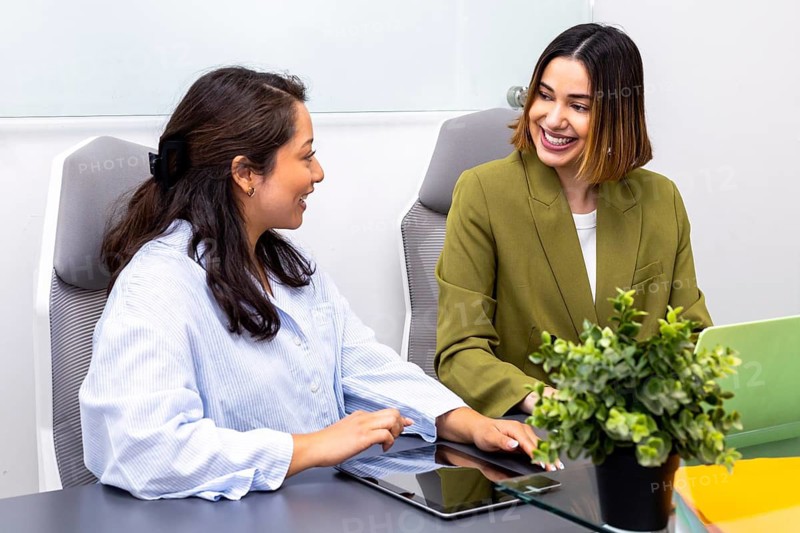 Two women engaged in a professional image consulting meeting, discussing strategies around a table with digital devices. A plant adds a touch of greenery to the office setting