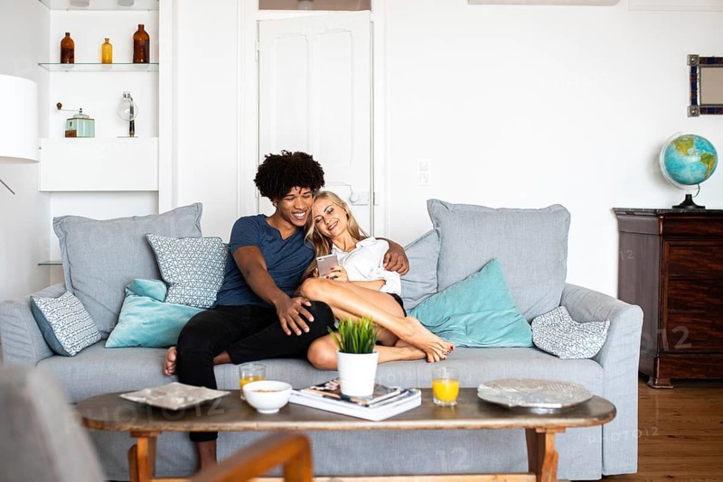 A happy couple sits on a cozy couch, looking at a phone. The bright living room has modern decor, with a globe on a nearby table, creating a relaxed atmosphere