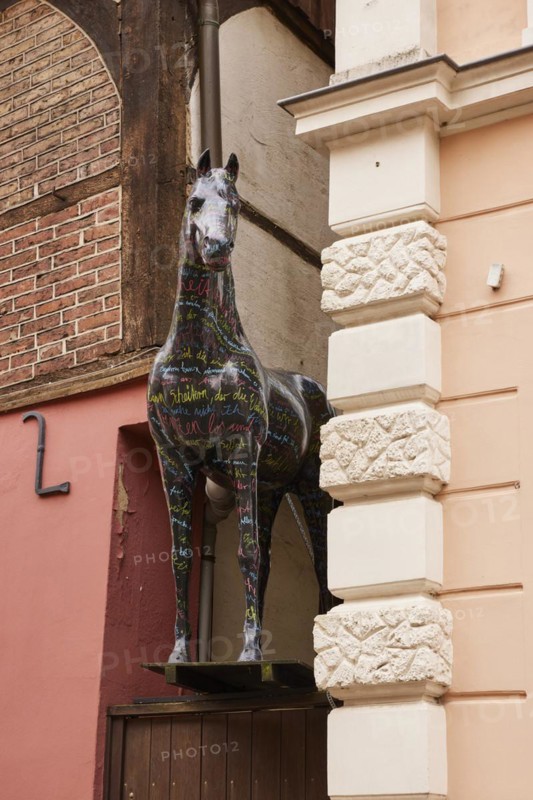 Horse sculpture with colourful lettering looks between a narrow passage between two buildings in the city of horses Warendorf, district of Warendorf, North Rhine-Westphalia, Germany