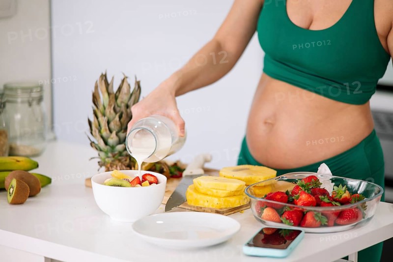 A pregnant woman in a green yoga outfit prepares a healthy breakfast with fresh fruits and milk. The setting emphasizes wellness and nutritious eating during pregnancy