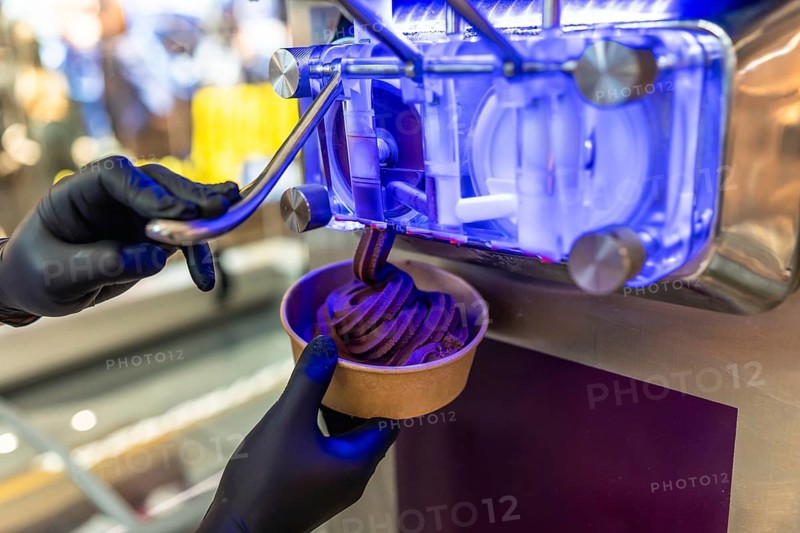 Person wearing black gloves operating a soft serve machine, dispensing delicious dark chocolate frozen yogurt into a paper cup, conveying a sweet dessert experience
