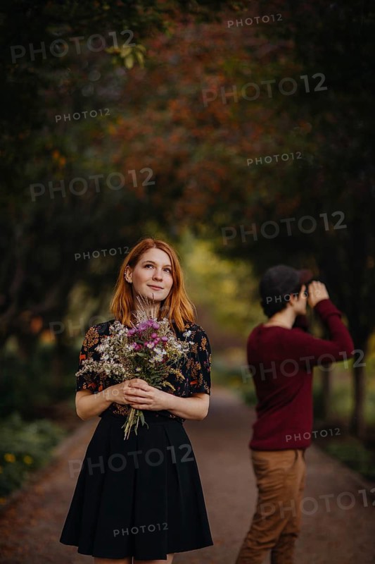 A woman holding a bouquet of wildflowers stands thoughtfully under colorful autumn trees, while a man enjoys the scenery with binoculars in a park in Quebec, Canada