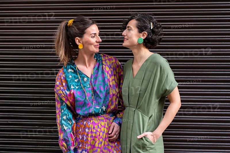 Two stylish women stand in cheerful clothing, gazing at each other with smiles. They are wearing colorful dresses and bold earrings in a vibrant street setting
