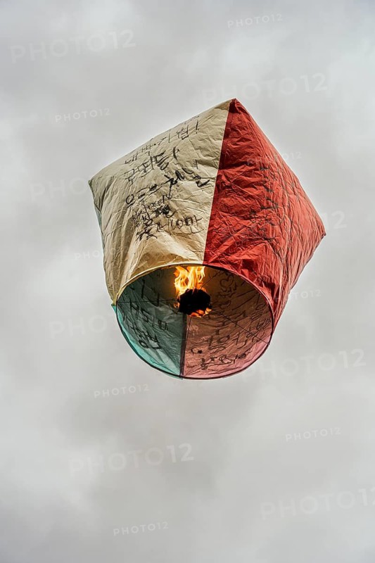 From below, this vibrant Chinese lantern ascends into an overcast sky, its surface covered with handwritten messages of health, happiness, and prosperity. Symbolic of traditional Taiwanese festivals, the lantern is seen as a bearer of personal hopes
