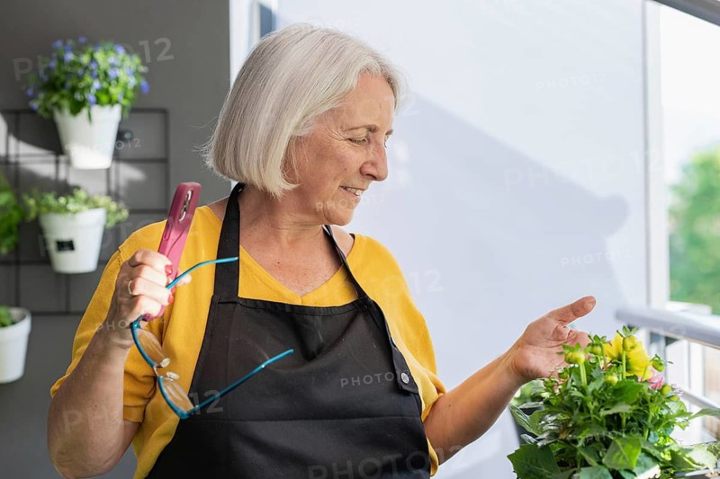 An elderly woman stands on a balcony, engaged with vibrant plants. She holds a smartphone and glasses, wearing a yellow shirt and black apron, enjoying gardening
