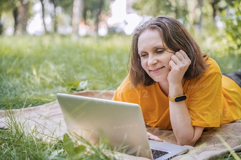 A woman is lying on a blanket outdoors in a park, using her laptop for work. She enjoys the serene environment, representing the modern trend of remote working