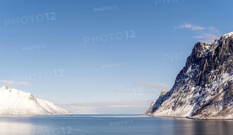 A breathtaking view of the snow-covered mountains and calm waters of the fjord in Mefjordbotn, Norway, under a clear blue sky, showcasing the serene beauty of winter