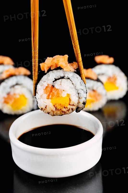 Close up of a sushi roll with salmon and mango, held by bamboo chopsticks above a bowl of soy sauce in black background. Perfect for culinary or Japanese cuisine themed content