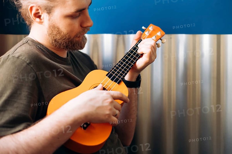 A musician with a beard passionately plays a bright orange Mexican ukulele, focused on his music. The background includes a blue wall and metal paneling, creating a modern look