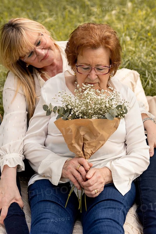 A serene moment capturing an elderly woman holding a bouquet while being lovingly embraced by her adult daughter. They are sitting outdoors, sharing a tender family bond