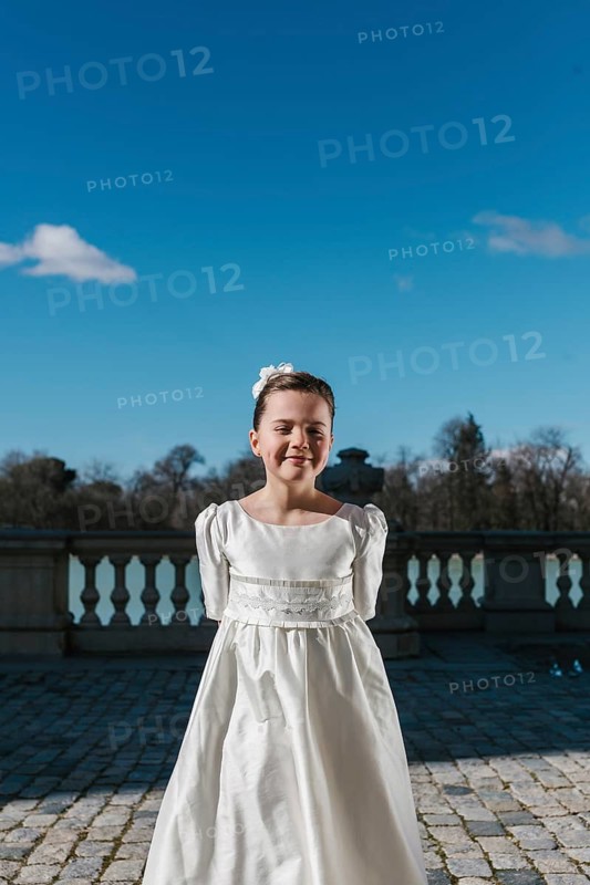 A young girl stands confidently in her white First Communion dress, under a clear blue sky. The setting is an open outdoor space with a serene backdrop