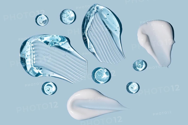 Various clear gel and white cream textures smeared and dotted on a pastel blue surface, showcasing beauty product textures for cosmetic or skincare-related content