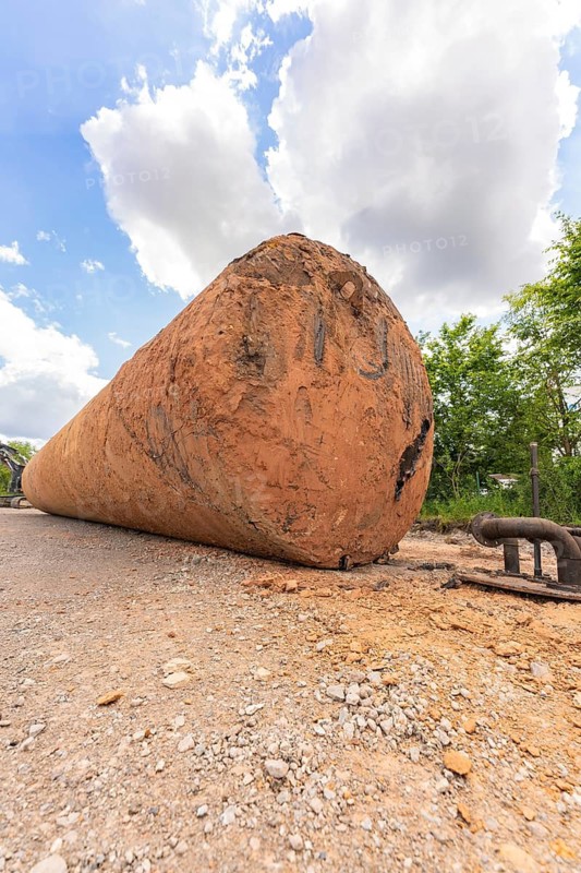 A large, brown tank lies flat on a construction site, surrounded by sky, clouds and trees, demolition site of a former lorry transport company with oil tanks, Saarbrücken, Germany