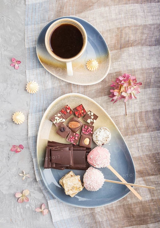 A pieces of homemade chocolate with coconut candies and a cup of coffee on a gray concrete background and blue and brown textile. top view, flat lay, close up