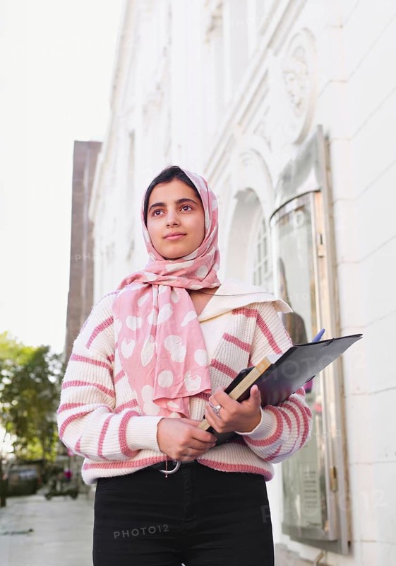 A arab student woman wearing a pink hijab stands outdoors, holding books and a clipboard. She is near a historic white building, looking forward with purpose