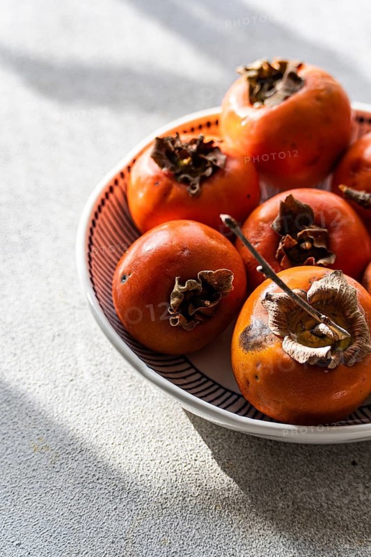 A bowl filled with freshly picked, ripe persimmons, positioned on a light textile surface, bathing in soft sunlight, highlighting their vibrant orange hue and soft textures