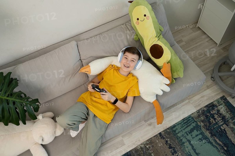 A happy boy wearing headphones plays video games while lounging on a cozy sofa. Surrounded by plush toys, he enjoys a relaxed gaming session at home