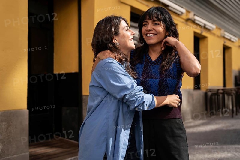 A happy multiethnic lesbian couple embraces, sharing a laugh in an urban environment. The moment captures love, pride, and diversity in a sunny setting