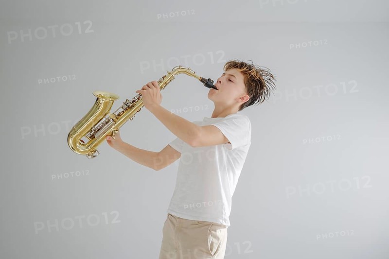 A young musician in a white shirt and beige pants passionately plays a golden saxophone. The minimalist background emphasizes the focus on music and expression