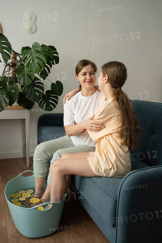 A mother and daughter share a serene moment on a sofa, relaxing with a foot soak filled with citrus slices. The cozy home environment fosters bonding and self-care