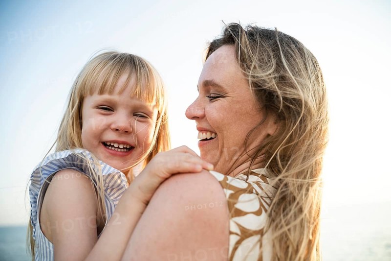 A cheerful mother and daughter share a joyful moment on a sunlit beach in Italy. The warmth of their smiles reflects the perfect family day by the seaside