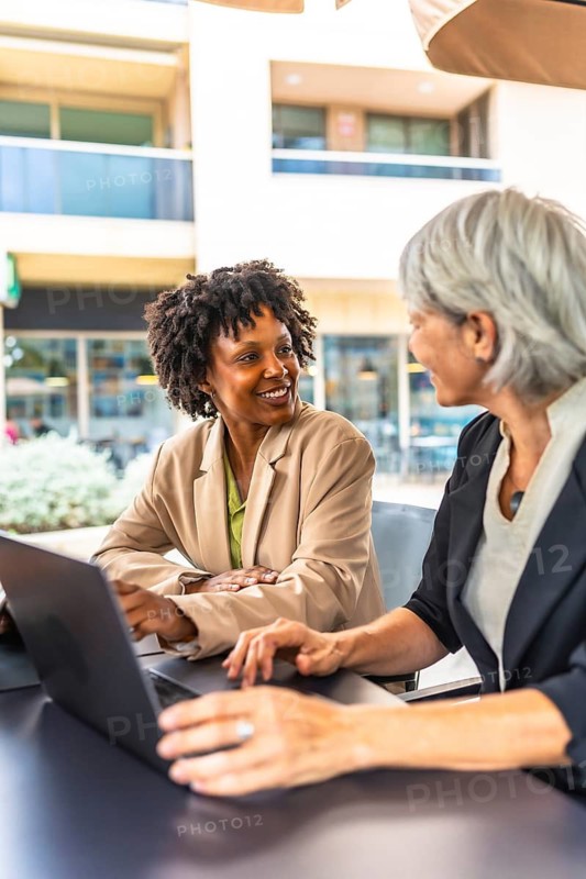Vertical photo of two multi-ethnic businesswoman working with laptop in a sidewalk cafeteria in the city