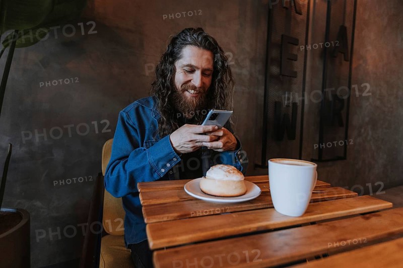 A man with long hair and a beard smiles while using his smartphone at a wooden table in a cozy cafe. A cup of coffee and a pastry are in front of him, creating a warm ambiance