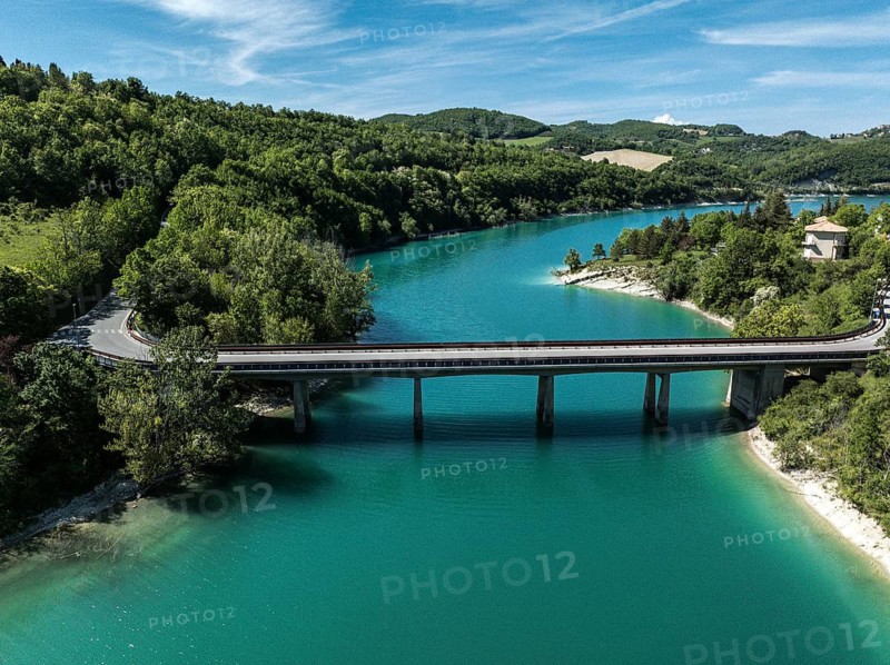Aerial view of a bridge spanning the vibrant turquoise waters of Lago di Fiastra, surrounded by lush green forests under a clear blue sky in Italy. Perfect for nature lovers