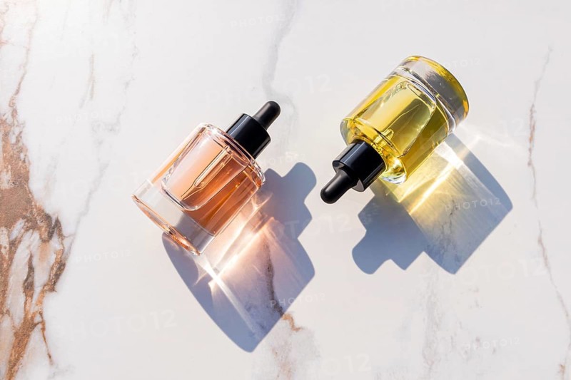 Two transparent skincare bottles with black droppers, filled with pink and yellow oils, sit on a marbled surface. Sunlight casts soft shadows, enhancing a minimalist aesthetic
