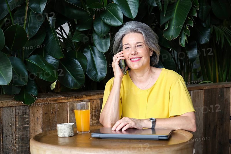 Mature woman, smiling in a yellow blouse, enjoys a phone call while sitting at a wooden table with a laptop and a glass of orange juice, surrounded by lush greenery