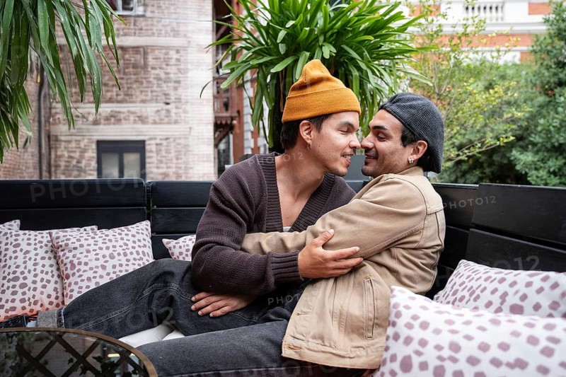 A loving Colombian gay couple sits closely on a cozy outdoor sofa surrounded by plants and pillows, sharing an affectionate moment in an urban setting