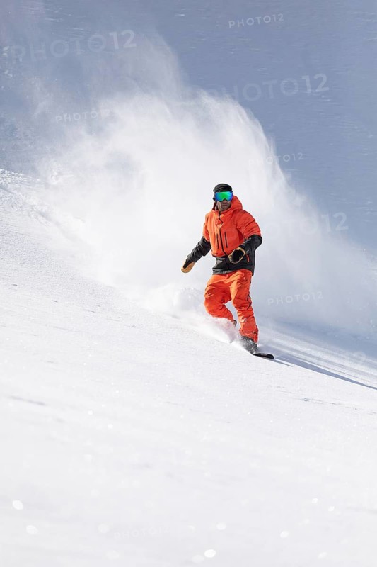 A snowboarder wearing bright orange gear rides down a snowy slope, leaving a trail of snow spray. The scene captures the thrill and excitement of winter sports action