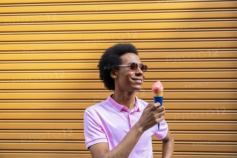A joyful non-binary person in sunglasses and pink shirt savoring an ice cream cone on a warm day, standing against a vibrant yellow background, embodying summer bliss